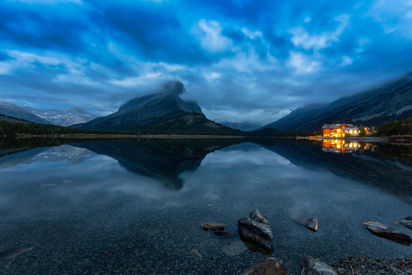 A night scene featuring a lakeside lodge with its lights reflecting on the still water, set against a backdrop of mountains and a twilight sky